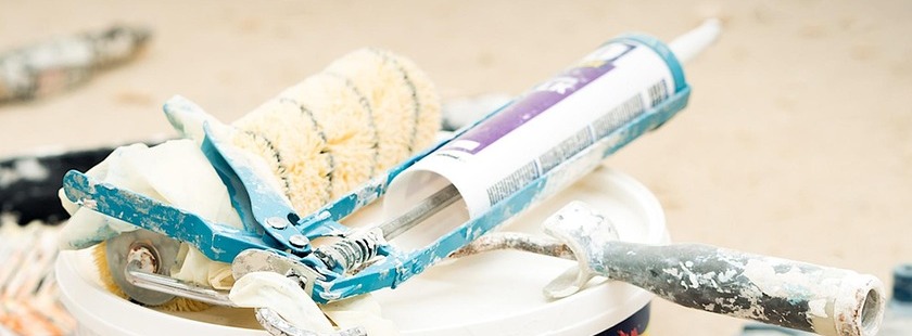Caulking gun loaded with a tube of sealant resting on a bucket lid beside a paint roller